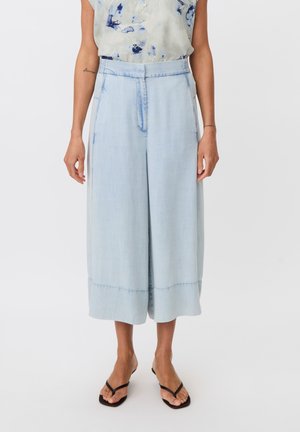 Mujer con pantalones vaqueros anchos cortos de color azul claro con cintura alta y sandalias negras de tiras, de pie frente a un fondo blanco.