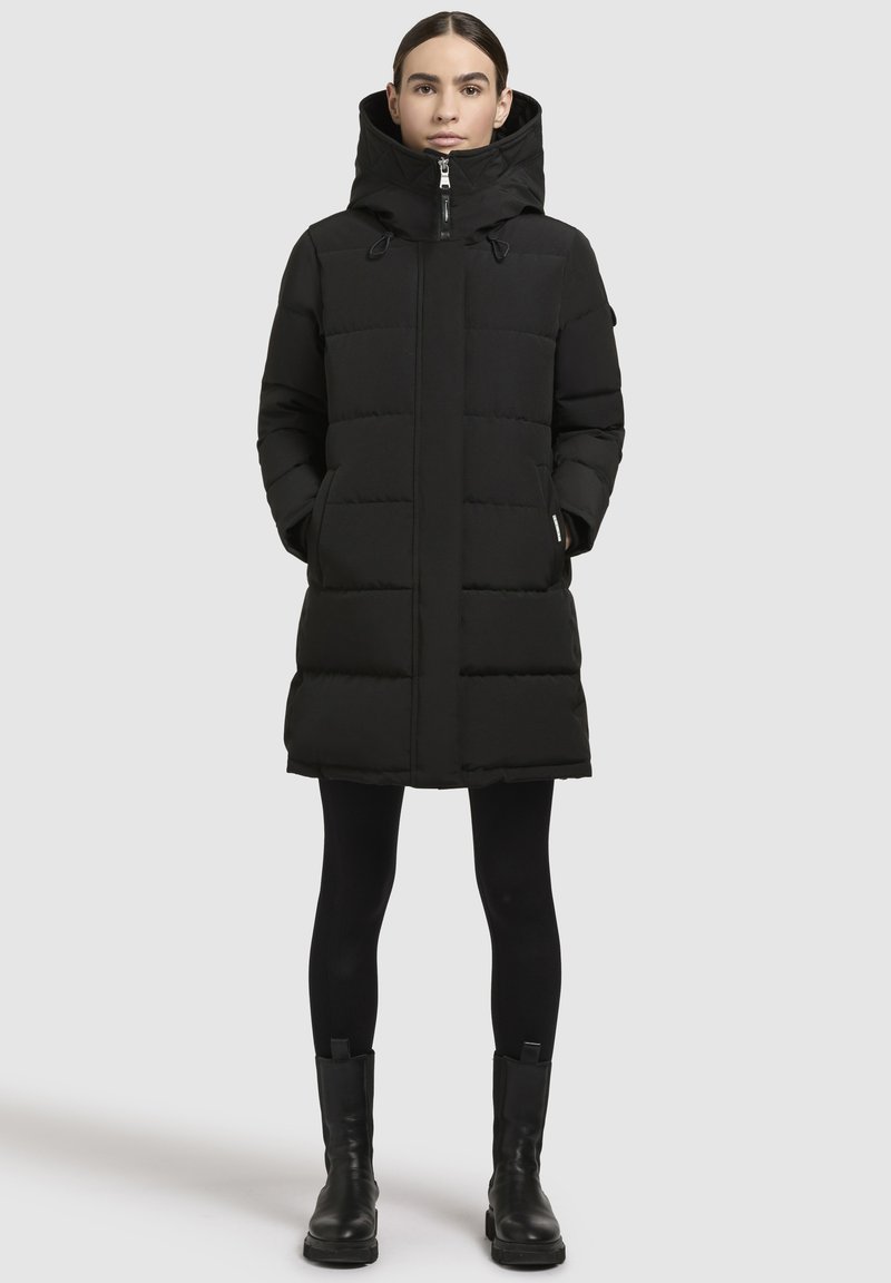 Sieviete, tērpta melnā kapucēta pūkainā mētelī, melnās legingās un melnās zābakos, stāv pretī vienkāršai gaišai fona.