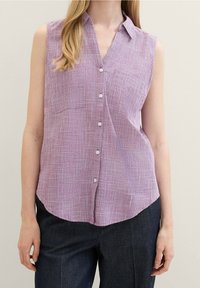 Camisa sin mangas de botones en púrpura claro con un patrón de cuadros texturizados, dobladillo curvado y dos bolsillos delanteros. Combinada con pantalones de mezclilla oscuros.