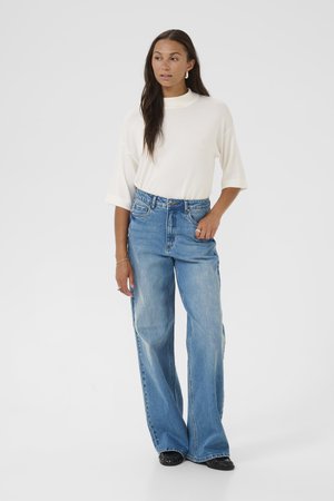 Woman with long brown hair wearing a white high-neck top with elbow sleeves, light blue wide-leg jeans, and black shoes, standing with hand in pocket.
