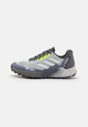 Hellgrauer und dunkelgrauer Adidas Terrex Trailrunning-Schuh mit weißen Streifen, schwarzen Schnürsenkeln, gelben Ösen und robuster Sohle, Seitenansicht.