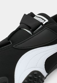 Gros plan sur une basket Puma noire et blanche avec un tissu texturé, une large bride et des crampons en caoutchouc sous la semelle.