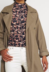 Beige trenčkot s knoflíkov ými detaily, přeložený přes tmavý květinový top, spárovaný s bílými džíny. Významné detaily zahrnují vysoký límec.