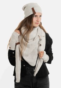 Lichtgrijze gebreide beanie en een oversized driehoekige sjaal, getextureerd met een subtiel patroon. Gecombineerd met vingerloze handschoenen, allemaal met bruine accenten.