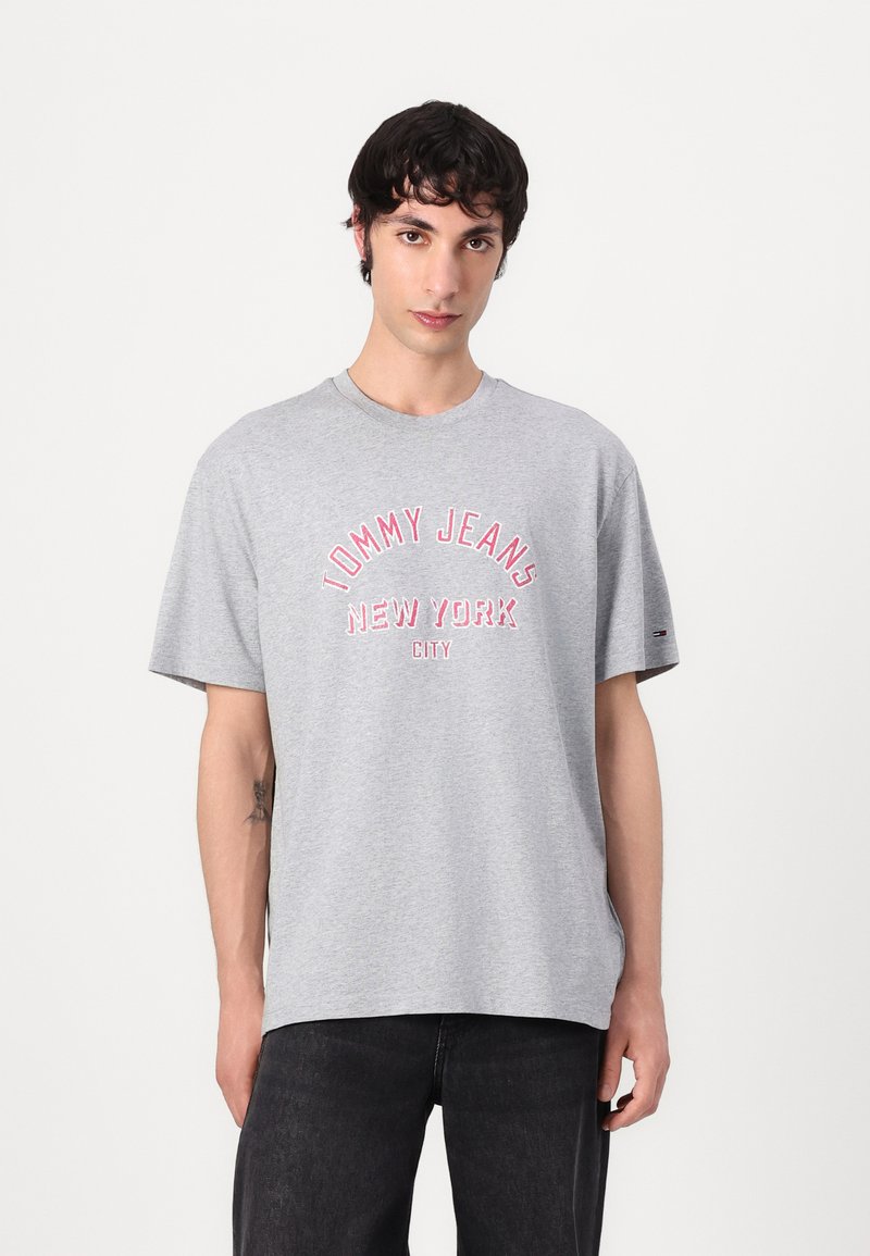 Grå bomulls t-skjorte med kort erm, med røde "TOMMY JEANS NEW YORK CITY"-tekst. Løs passform, rund hals, og en liten logo på ermet.