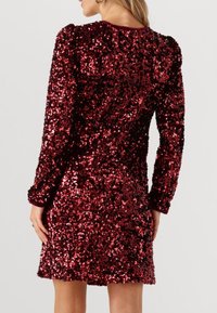 Robe rouge en sequins avec manches longues, dotée d'une silhouette ajustée et d'une texture brillante, reflétant la lumière avec de petits sequins circulaires.