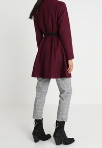 Bordový kabát s páskem a dlouhými rukávy, spárovaný se šedými kostkovanými kalhotami a černými kotníkovými botami na podpatku se stříbrnými kulatými akcenty.