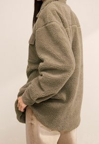 Fuzzy olijfgroene fleece jack met een kraag, geribbelde manchetten en een borstzak, gedragen over licht tan kleurige jeans.