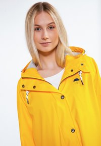 Gelbe wasserdichte Jacke mit einem grauen Kragen, Frontreißverschluss und Druckknöpfen. Mit einem Regenschirm-Logo und Kordelzügen.