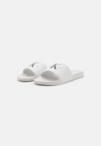 Hvite sklisikre sandaler med teksturert overdel som har en svart "CK" logo. Myk såle med flat design, minimalistisk og lett.