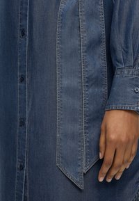 Τζιν πουκάμισο με κάθετες ραμμένες πλάκες και μανσέτες με κουμπιά, εμφανίζεται στον βραχίονα και το χέρι ενός ατόμου που ακουμπούν στο πλάι του.