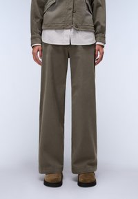 Καμηλό παντελόνι με φαρδιά γραμμή φτιαγμένο από λείο ύφασμα, σε συνδυασμό με ένα ανοιχτό ριγέ πουκάμισο και καφέ σουέντ παπούτσια χωρίς λουριά.