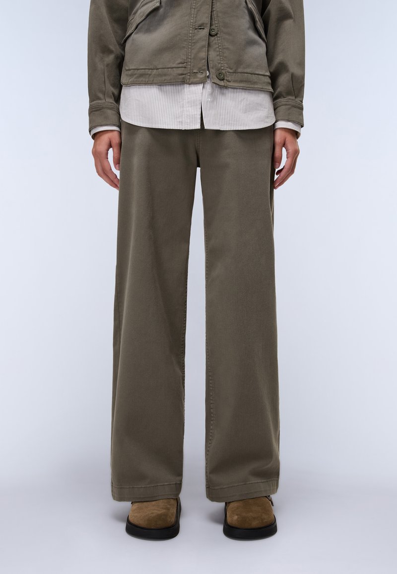 Καμηλό παντελόνι με φαρδιά γραμμή φτιαγμένο από λείο ύφασμα, σε συνδυασμό με ένα ανοιχτό ριγέ πουκάμισο και καφέ σουέντ παπούτσια χωρίς λουριά.