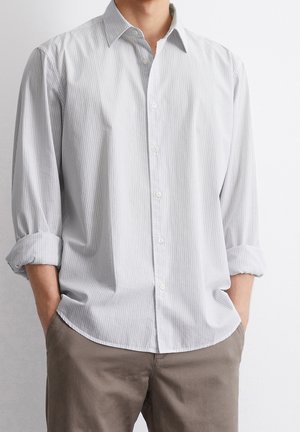 Homme portant une chemise gris clair à manches longues avec les manches retroussées et un pantalon marron, mains dans les poches sur un fond uni.
