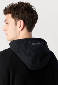 Črna kapuca z teksturirano tkanino in gladko kapuco. Notranja kapuca prikazuje natisnjen tekst. Posnetek od blizu zadaj.