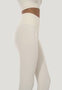 Kvinderkrop og ben iført sømløse, højtaljede cremefarvede leggings og en matchende crop top mod en ensfarvet baggrund.