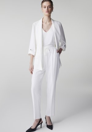 Mujer vestida con blazer blanco, camiseta blanca de cuello en V, pantalones blancos con cordón y zapatos de tacón negro con tiras en el talón, posando frente a un fondo liso.