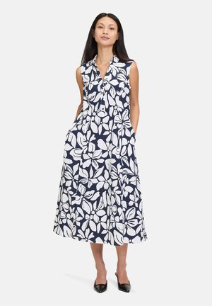Navyblaues, ärmelloses Kleid mit weißem Blumenmuster, kragenförmigem Design, Taschen, Midi-Länge und fließendem Stoff. Schwarze spitze Pumps.