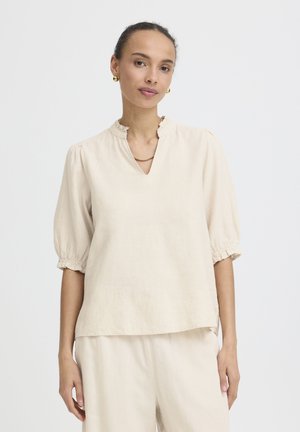 Žena nosi smotanú blúzku s nafúknutými rukávmi a zladené krémové nohavice, doplnené zlatými náušnicami a náhrdelníkom.