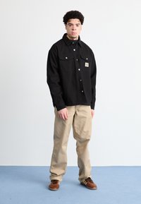 Sort skjorte med knapper og to brystlommer samt et logo-patch, parret med khaki bukser og brune sneakers. Ensfarvede nuancer, afslappet pasform.