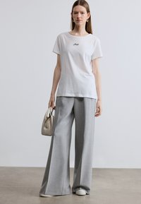 Λευκό κοντομάνικο μπλουζάκι με μαύρο κεντητό κείμενο, σε συνδυασμό με ανοιχτόγριες παντελόνες με φαρδιά μπατζάκια. Το μοντέλο κρατά μια μικρή μπεζ τσάντα.