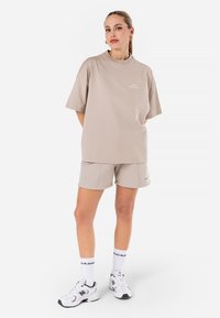 Beige bomulls t-skjorte med korte ermer og rund hals, kombinert med matchende shorts. Hvite joggesko med svarte detaljer og mønstrede sokker.