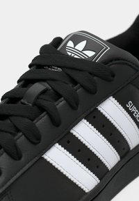 Black leather sneaker with black laces, featuring three white stripes and a branded black and white tongue label. Textured material details present.