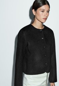 Black cropped wool jacket with round neckline, five black buttons, and long sleeves. Smooth texture with a clean, minimal design.
