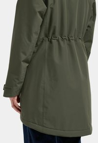 Olivgrön, lätt jacka med elastisk midja, slät yta och justerbara ärmslut. Längden är något rundad.