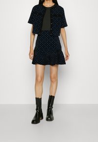 Femme portant une veste noire à manches courtes avec motifs et une jupe évasée assortie, un top noir en dessous, et des bottines noires à talons.