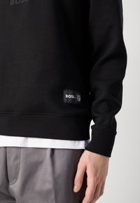 Sweatshirt preto feito de uma mistura de algodão, com punhos e barra canelados, e um rótulo de logo na parte inferior esquerda. Usado por cima de uma camisa branca.