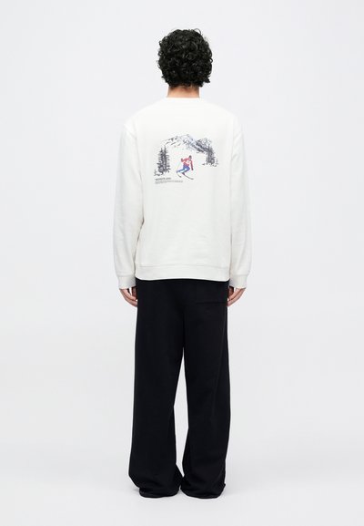 Krémová mikina s potlačeným motívom lyžiara a hôr na chrbte. Kombinovaná s voľnými čiernymi nohavicami.
