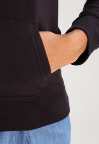 Sweat à capuche noir avec poche avant et poignets côtelés, associé à un pantalon bleu clair. Le tissu semble doux et légèrement texturé.