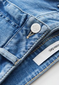 Jeans en denim bleu clair avec un bouton argenté portant un logo gravé, des coutures visibles et une étiquette en tissu. La texture est lisse et usée.