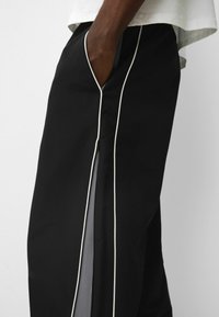 Persoon in zwarte broek met witte piping, hand in de zak, en een kleine zijrits nabij de knie, gecombineerd met een lichtgekleurde top.