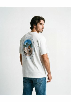 Hombre con cabello oscuro, vistiendo una camiseta blanca con un gran gráfico de un oso junto a un coche y palmeras, combinado con vaqueros azules, mirando de lado.