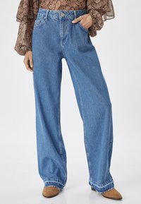 Jeans in denim blu con gamba larga e vita alta, tasche frontali e orlo sfilacciato. Il tessuto è leggermente sbiadito, offrendo un aspetto casual.