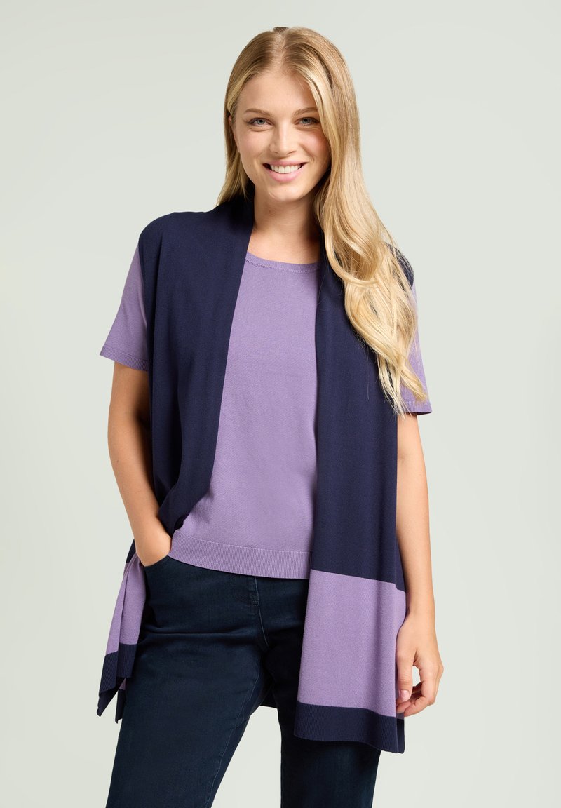 Chaleco sin mangas azul marino con un dobladillo lila sobre una camiseta lila. El conjunto presenta telas suaves y lisas con un corte relajado. Pantalones de mezclilla oscura.