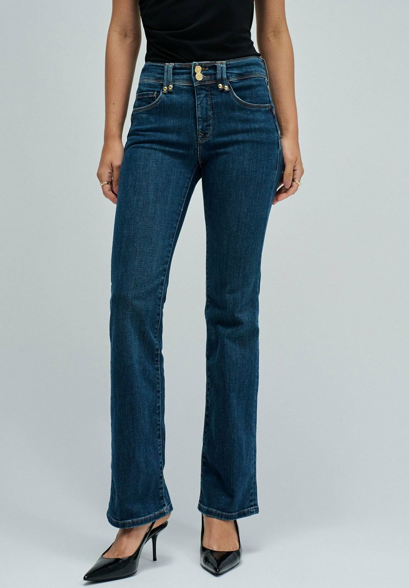 Jeans de tiro alto en azul oscuro con un corte ajustado, que cuentan con dos bolsillos delanteros y detalles en hardware dorado. Largo hasta el suelo con un ligero acampanado en el dobladillo.