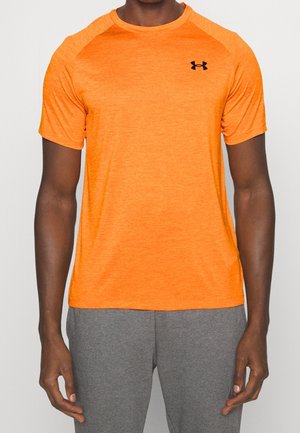 Mann trägt helloranges kurzärmeliges Sportshirt mit schwarzem Logo und grauer Sporthose vor einfarbigem Hintergrund.