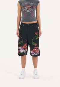 Shorts de mezclilla negra con patrones florales y texto en las piernas, combinados con una camiseta sin mangas gris ajustada con gráfico. Zapatillas blancas completan el look.