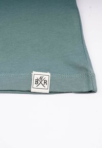 Helle grüne Baumwoll-T-Shirt mit glatter Textur, ausgestattet mit einem weißen Etikett mit schwarzer Schrift am Saum. Einfaches Design und einfarbig.