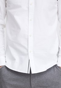 Hvid skjorte med knaplukning i glat stof, der har seks knapper, en buet kant og diskrete syninger nær sidesømmen.