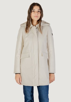 Femme portant un manteau léger beige à capuche avec poches à rabat, debout devant un fond uni, associée à un jean bleu.