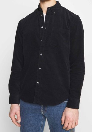 Mand iført sort kordfløjls-skjorte med knapper over hvid top og blå jeans, stående mod en ensartet lys baggrund.