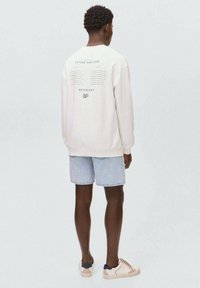 Sweatshirt crème avec un graphisme textuel au dos, associé à un short en denim bleu clair. Les bas sont effilochés à l'ourlet ; les chaussures sont beiges avec des accents bleu marine.
