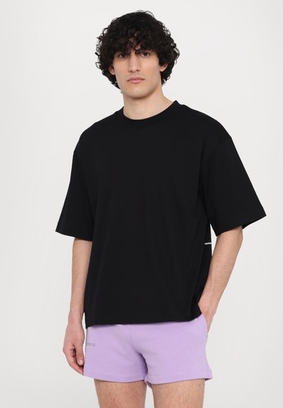 Camiseta de algodón negra con corte holgado, cuello redondo y mangas cortas, combinada con pantalones cortos lilas. Diseño sencillo, sin patrones visibles.