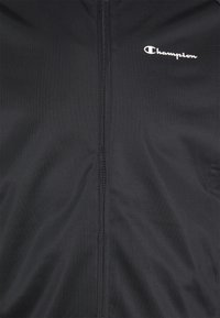 Tissu noir avec une fermeture éclair visible au centre et un logo blanc "Champion" en haut à droite.