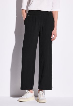 Personne portant un pantalon noir ample texturé, une chemise blanche à manches courtes et des baskets blanches, se tenant contre un mur blanc.