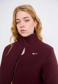 Raudonas Nike džemperis su aukštu apykakle, užtrauktuku priekyje ir baltu logotipu. Audinys atrodo lygus ir švelnus, su struktūrizuotu dizainu.